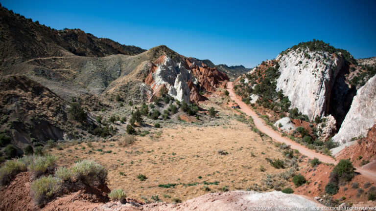 A dirt road crosses an open basin between pale cliffs and red ridges.