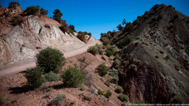 A dirt road bends between a pale fractured slope and a dark ridge, passing a rounded shrub.