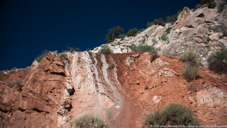 A steep red and white cliff shows vertical fractures and sparse shrubs along its rim.