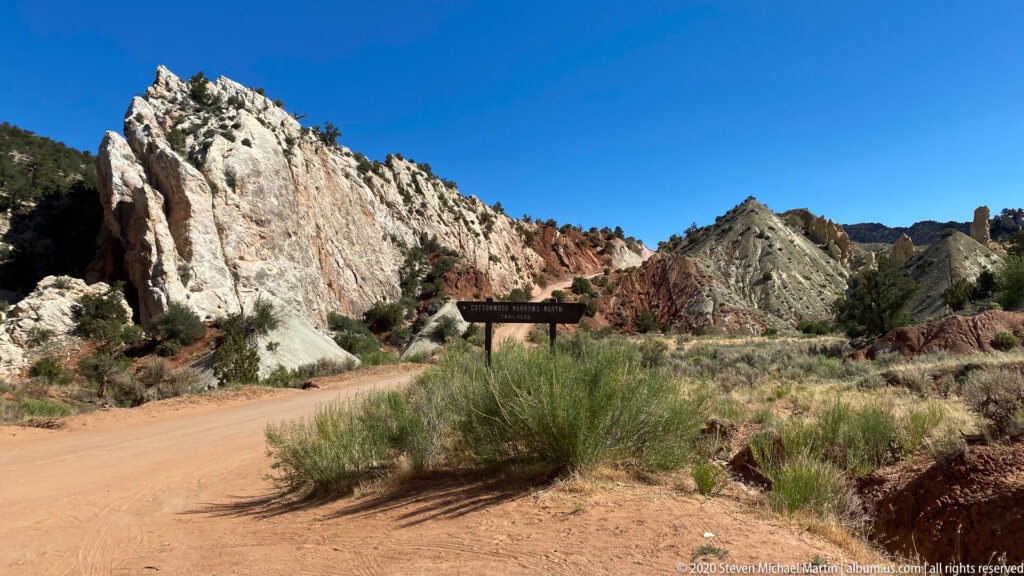 A Cottonwood Narrows sign stands beside a sandy road beneath pale and red rock formations.