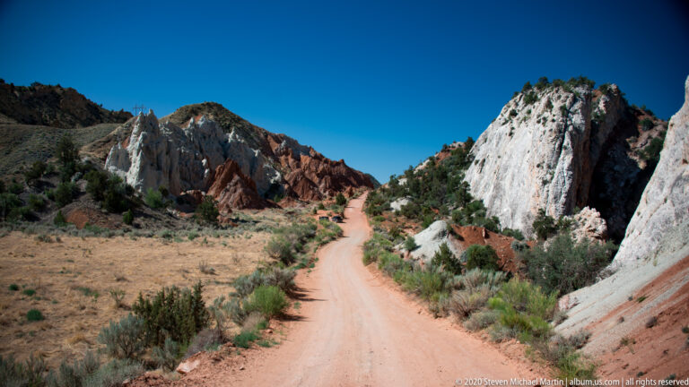 A sandy road leads between a tall white cliff and red-and-gray formations in the distance.