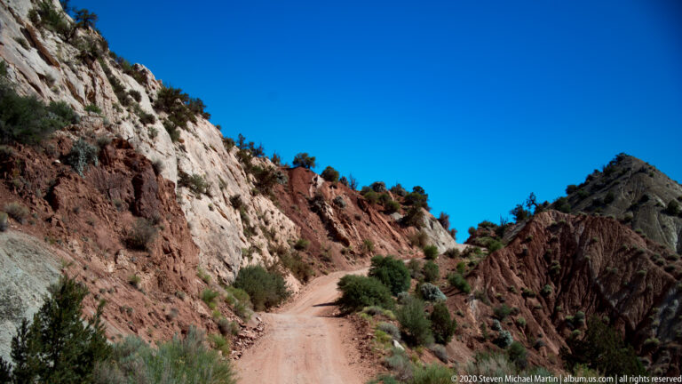 A dirt road curves between red and pale sandstone walls beneath a clear blue sky.