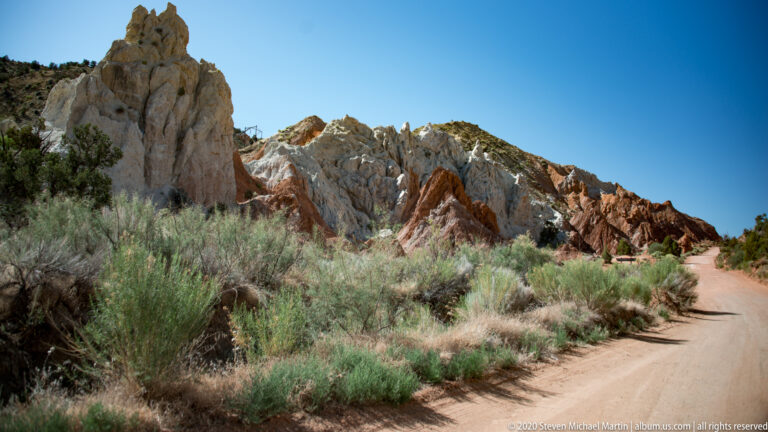 A dirt road curves past pale sandstone fins and rust-red ridges amid desert brush.