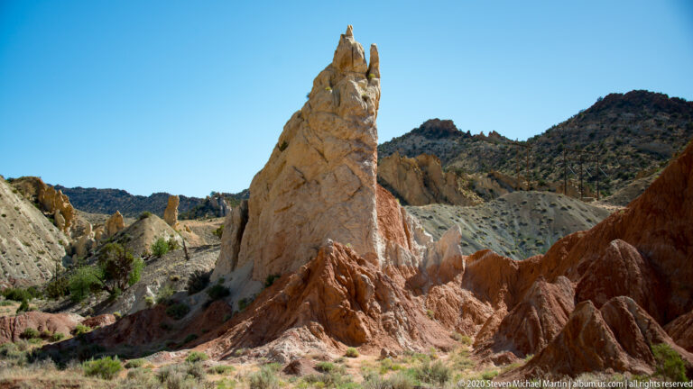 A pale sandstone spire rises from a red eroded slope before dark hills.