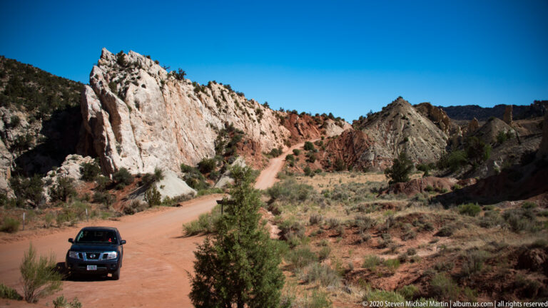 A dark SUV follows a dirt road through red and cream sandstone hills.