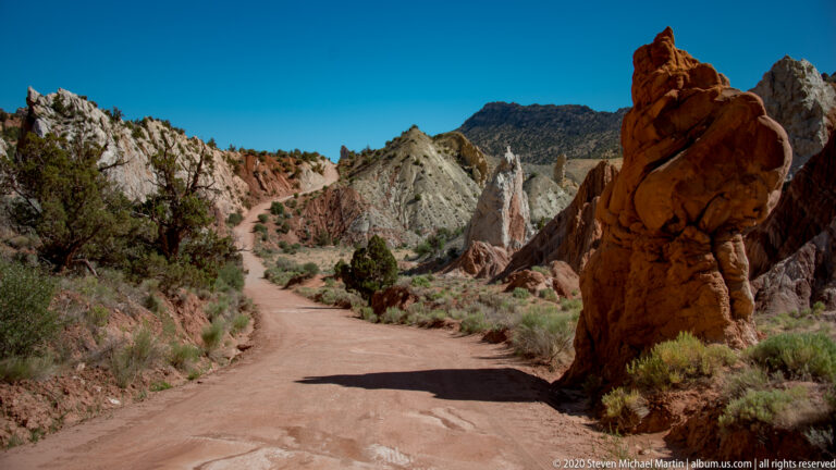 A dirt road runs between pale canyon walls and a large red sandstone outcrop.