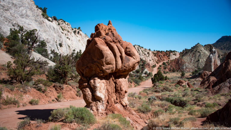 A bulbous red sandstone cap sits on a narrow pedestal beside a dirt road.
