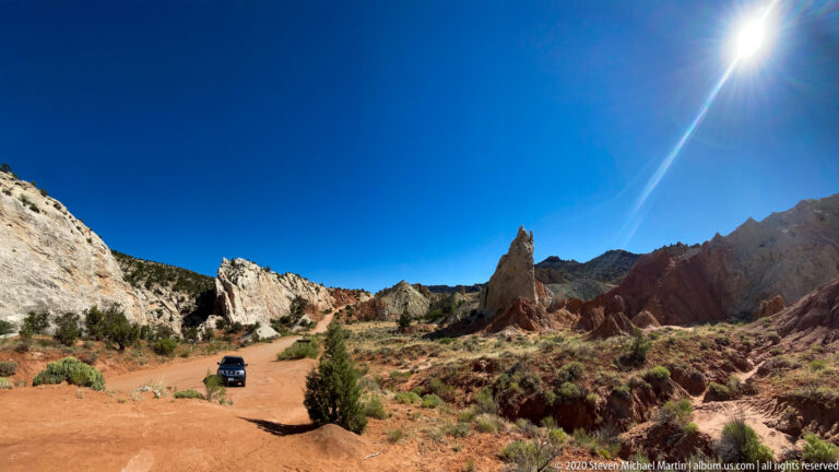 A dirt road and lone shrub lead toward a rock spire between pale and red cliffs under bright sun.