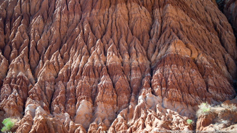 A steep close-up shows narrow erosion channels descending across a reddish sandstone slope with pale stone near its base.
