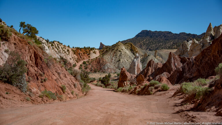 A dusty track winds between red fins and pale ridges beneath distant dark hills and a clear blue sky.
