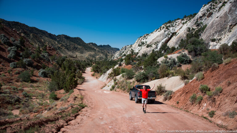 A visitor in red raises both arms beside an SUV on a dirt road between canyon walls.