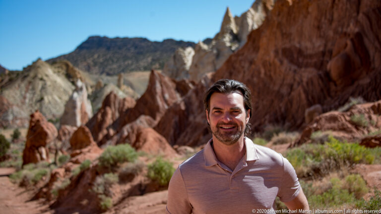 A man in a light polo shirt stands before softly blurred red sandstone formations.