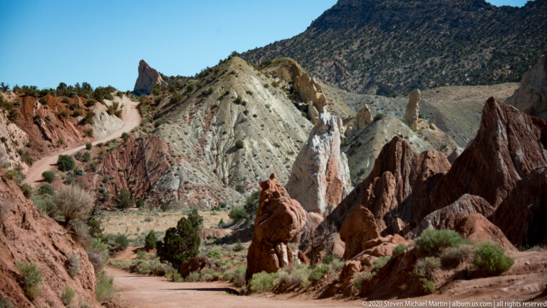 A dirt road curves between a pointed red rock and a pale eroded ridge.