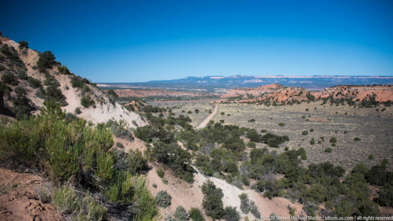 A pale rocky slope drops toward a straight dirt road crossing a broad shrub-covered valley.