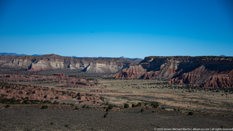 Sparse desert scrub fills a basin beneath distant layered gray, red, and cream cliffs.
