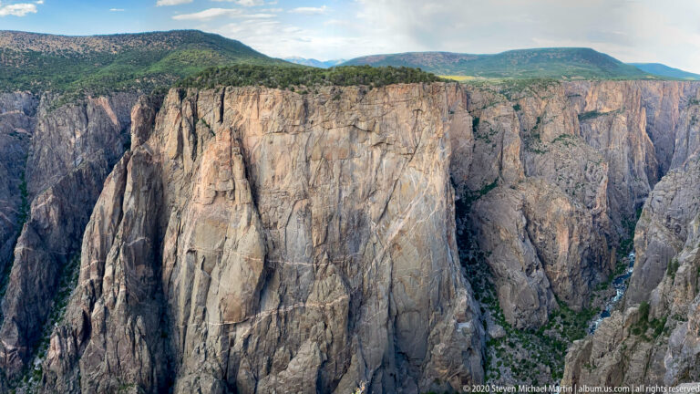Parallel fractures cross pale canyon walls above a narrow green strip deep below.