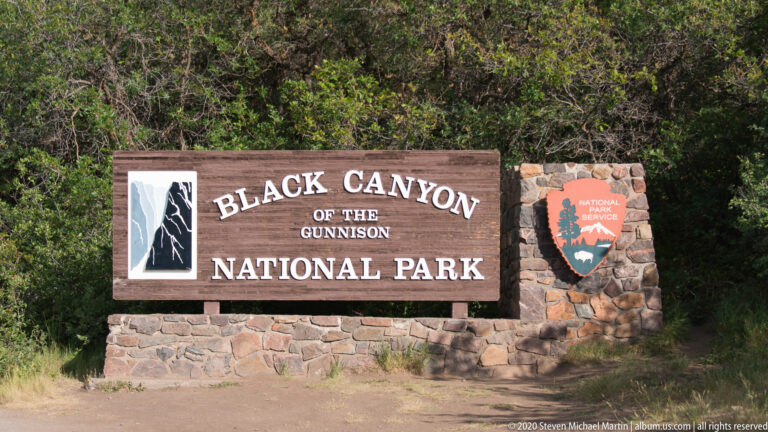 A wooden Black Canyon of the Gunnison National Park sign stands on a stone base beside an NPS shield.