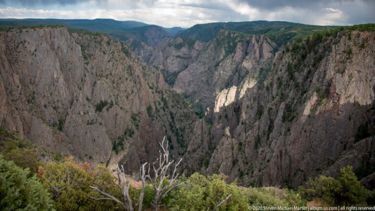 A sunlit patch marks one far cliff above a deep gorge framed by shrubs and a bare branch.