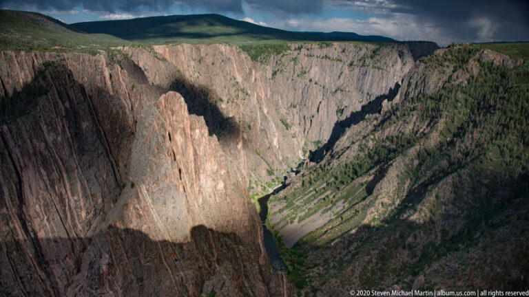 A sunlit cliff contrasts with shadowed walls above a distant river and green ravine.