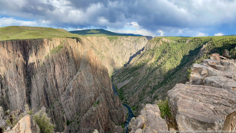 A grassy rim overlooks a broad cliff face and the winding Gunnison River below.