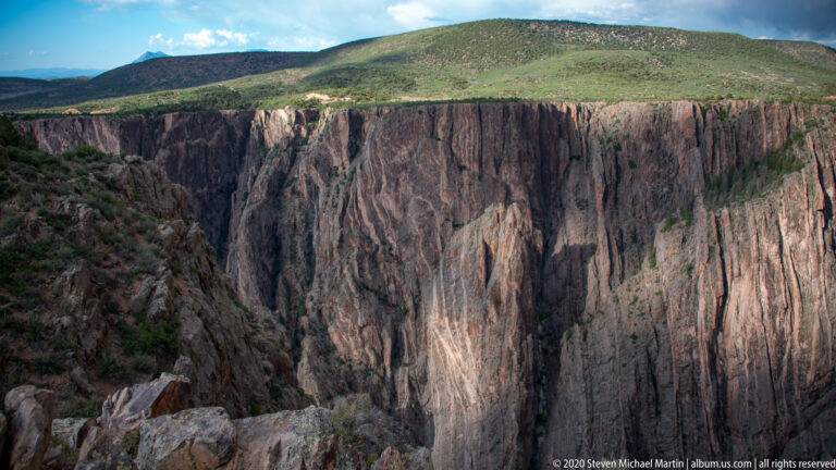 A fractured canyon wall rises from foreground boulders to a grassy plateau.