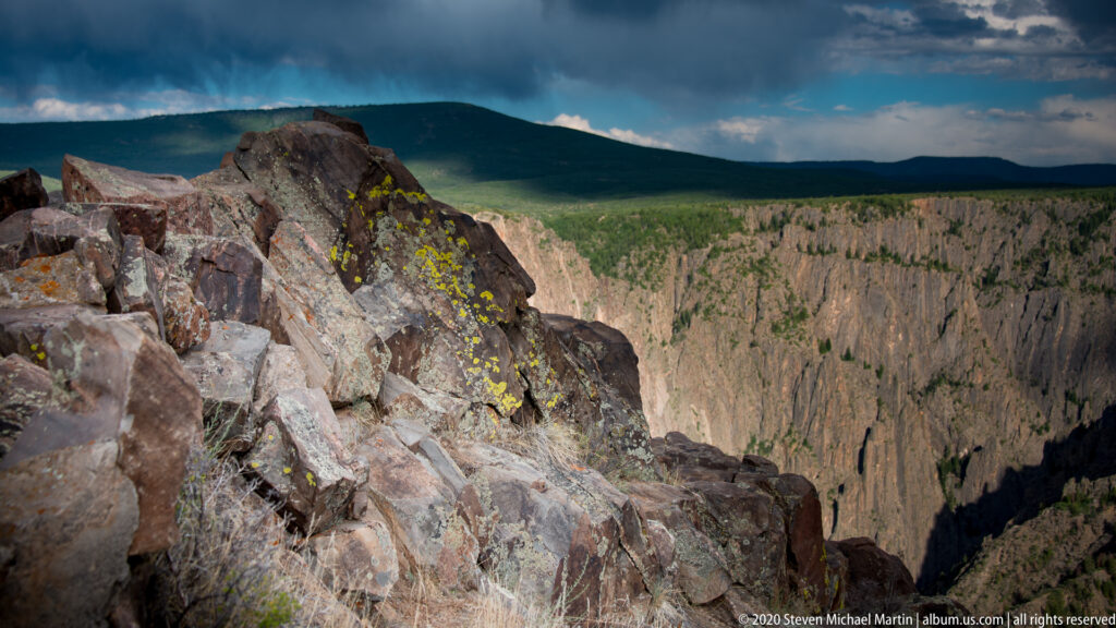 Lichen-covered boulders frame a steep canyon drop beneath a distant green horizon.