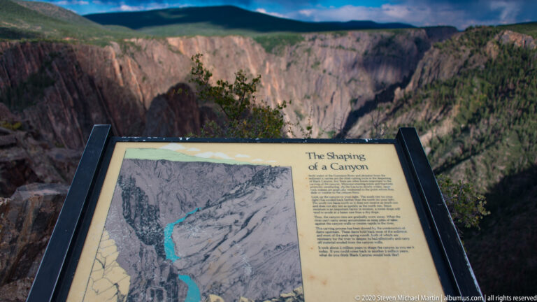An interpretive sign stands at the rim with the canyon cliff behind it.