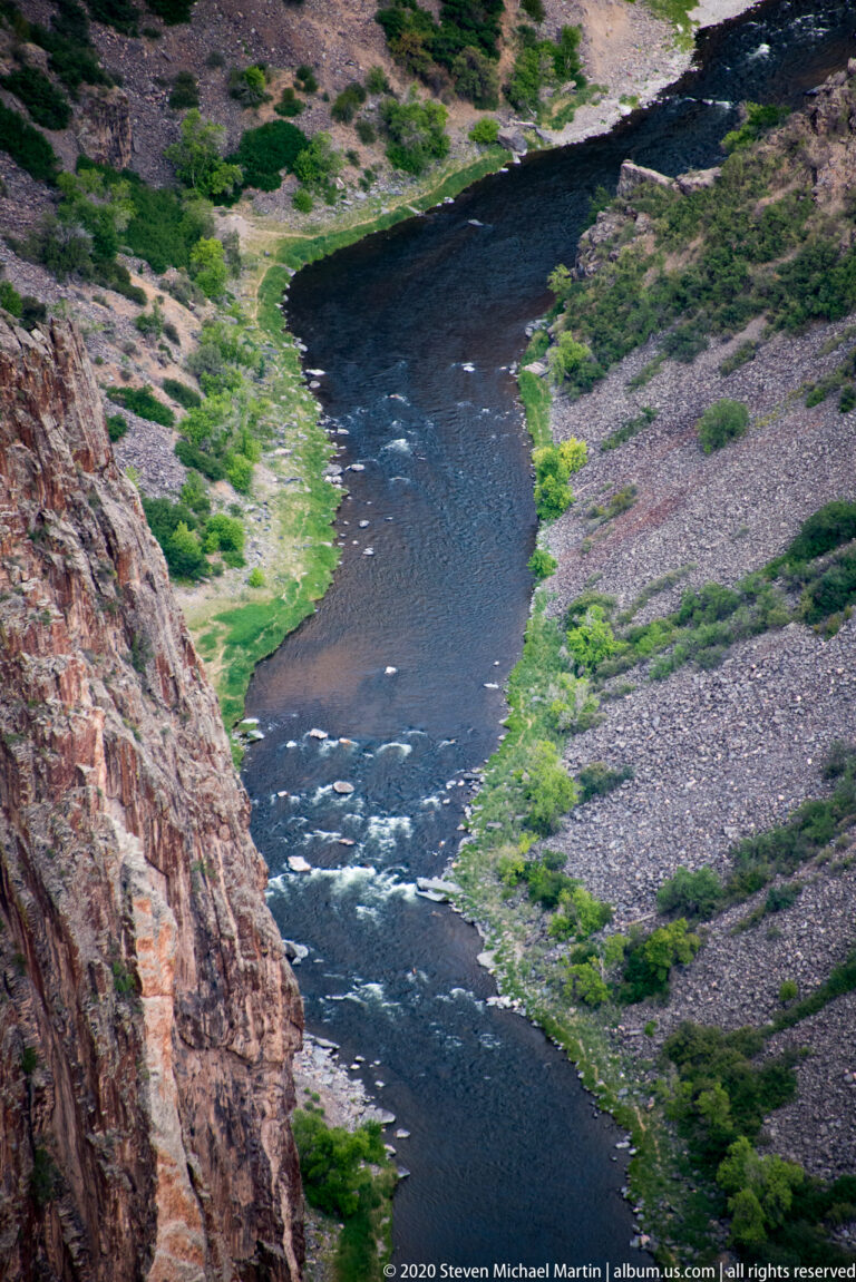 The Gunnison River winds around gravel bars and green patches below steep canyon slopes.
