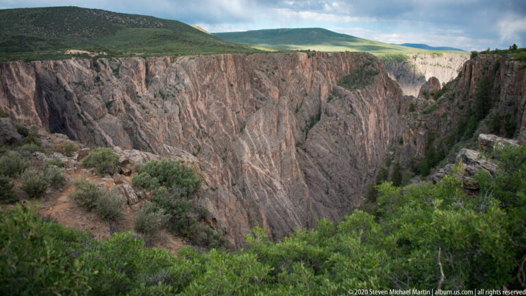 A green plateau meets a banded cliff that descends toward a vegetated river corridor.