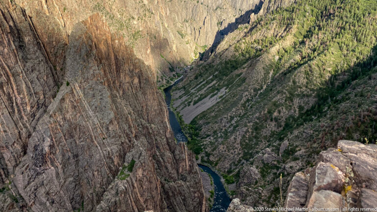 A small river bend lies far below between steep, vertically striated canyon walls.