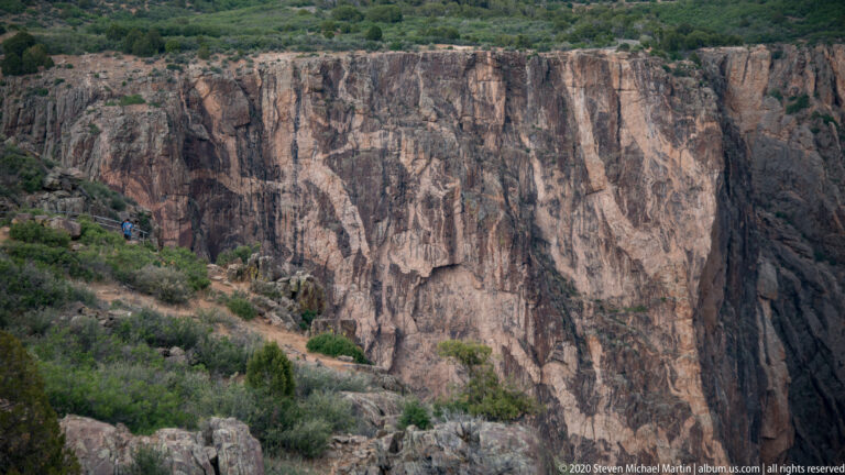 Two visitors walk a narrow trail beneath an immense fractured canyon wall.