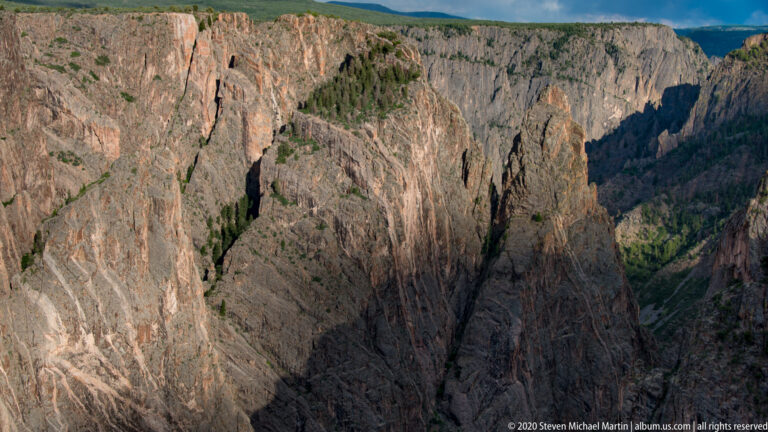 Fractured canyon walls drop toward a dark river gorge, with scattered evergreens on the ledges.
