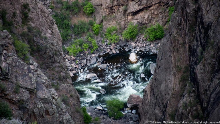 Whitewater rushes around boulders in the Gunnison River beneath steep, shrub-lined canyon walls.
