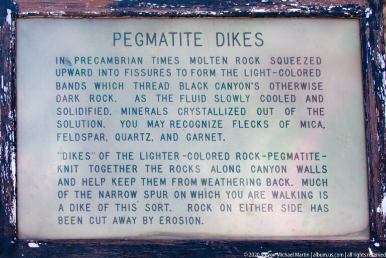 An interpretive sign describes light-colored pegmatite dikes cutting through dark rock in the canyon.
