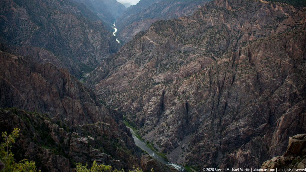 A pale river winds far below between dark, forested slopes of a deep canyon.
