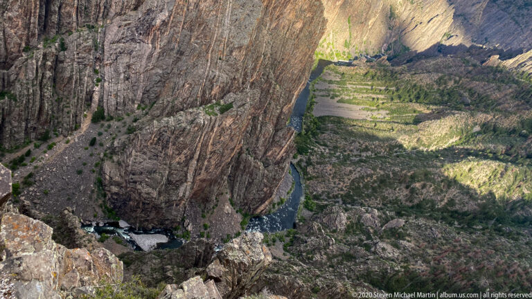 A high view looks down to the Gunnison River bending around gravel bars between sheer cliffs.