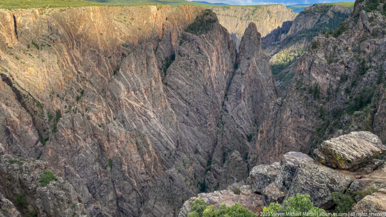 Banded canyon cliffs descend toward a partly hidden river, seen across foreground boulders.