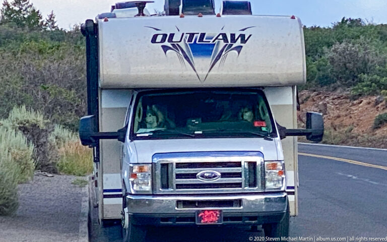 A white motorhome approaches on a paved road bordered by sagebrush and low trees.
