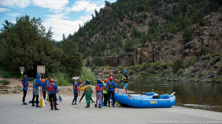 Helmeted paddlers gather with a blue raft and paddles beside the river.