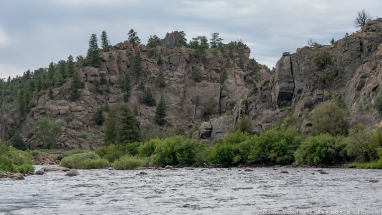 A broad river channel lies below a pine-covered rocky slope and green shrubs.