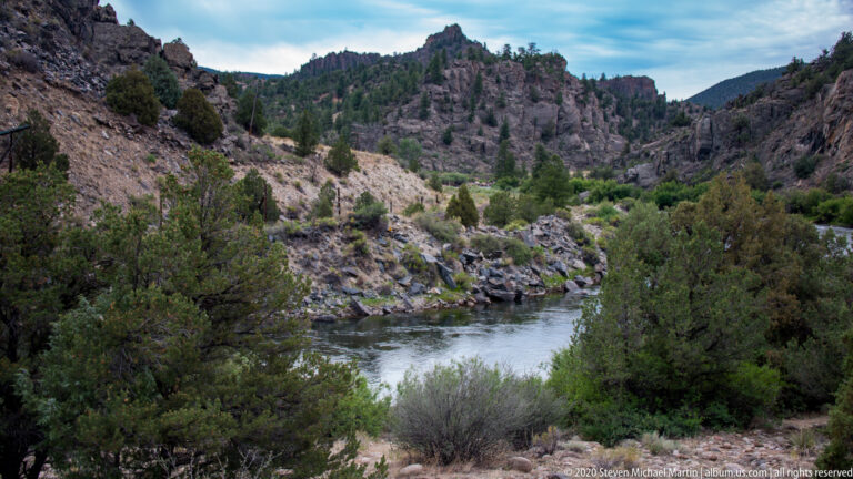 A river winds through the valley between boulder-strewn slopes dotted with pines and shrubs.