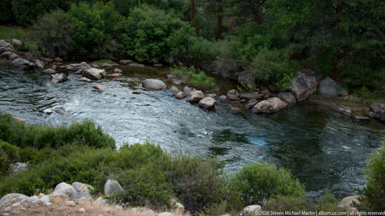 A river rapid flows around rounded boulders between dense shrubs and conifers.