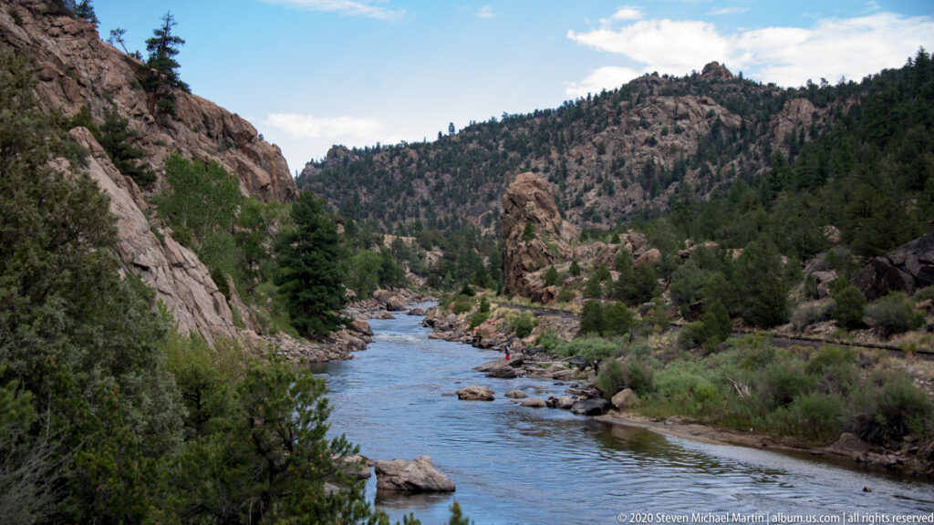 A small raft with a red-clad paddler moves through a rocky river corridor.