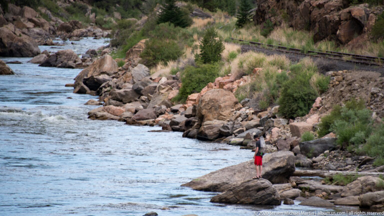 A person in red shorts stands on a boulder beside a flowing river.