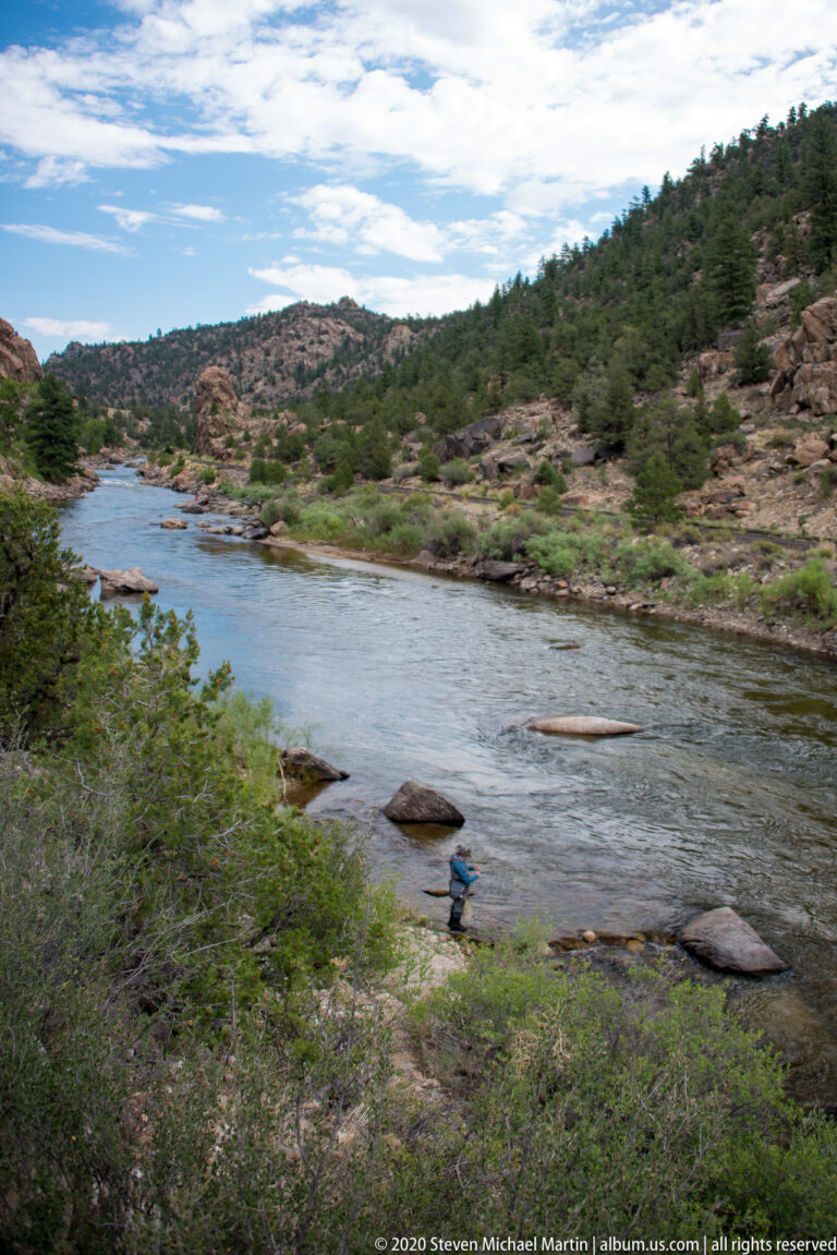 A person in blue sits beside a broad river channel partly screened by shrubs.
