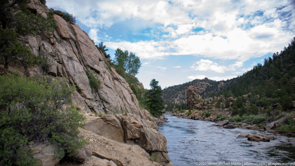 A river flows below a steep rock face and pine-covered canyon slopes.