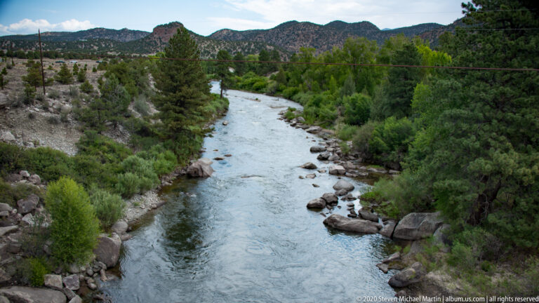 A river runs through a wooded valley beneath distant rocky mountain ridges.