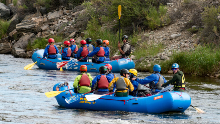 Two rafts of helmeted paddlers pass a guide standing on the rocky riverbank.