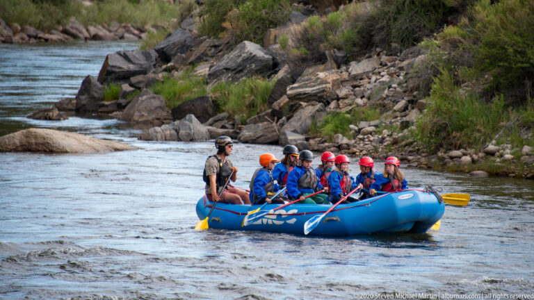 A blue raft carries helmeted paddlers through a shallow channel between rocky banks.