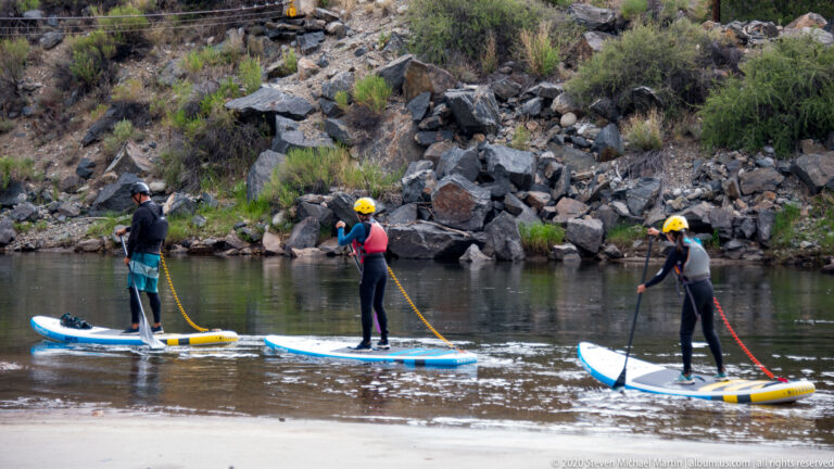 Three helmeted people stand on paddleboards and use long paddles on the river.